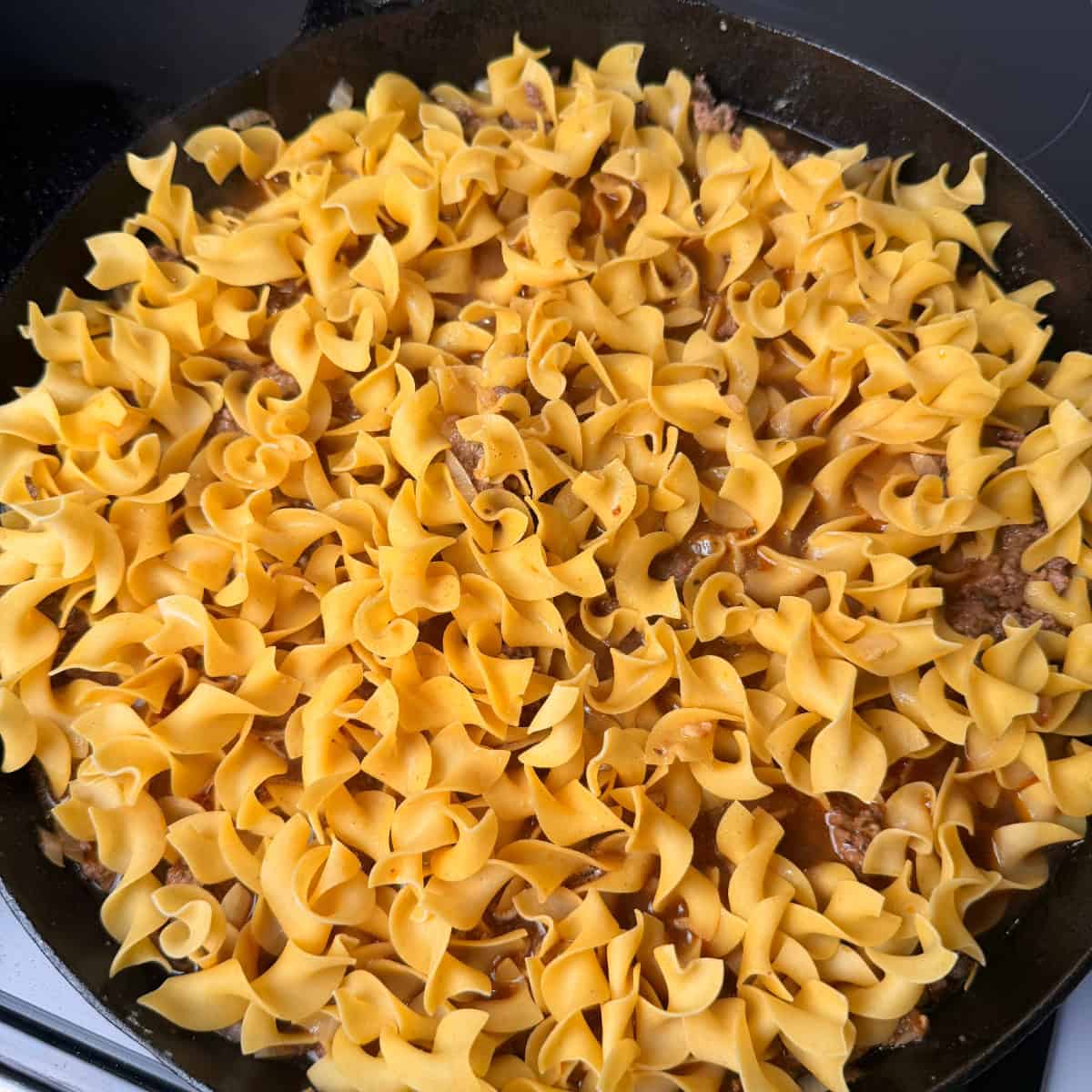 Beef, seasonings, onions, and egg noodles in skillet with beef broth