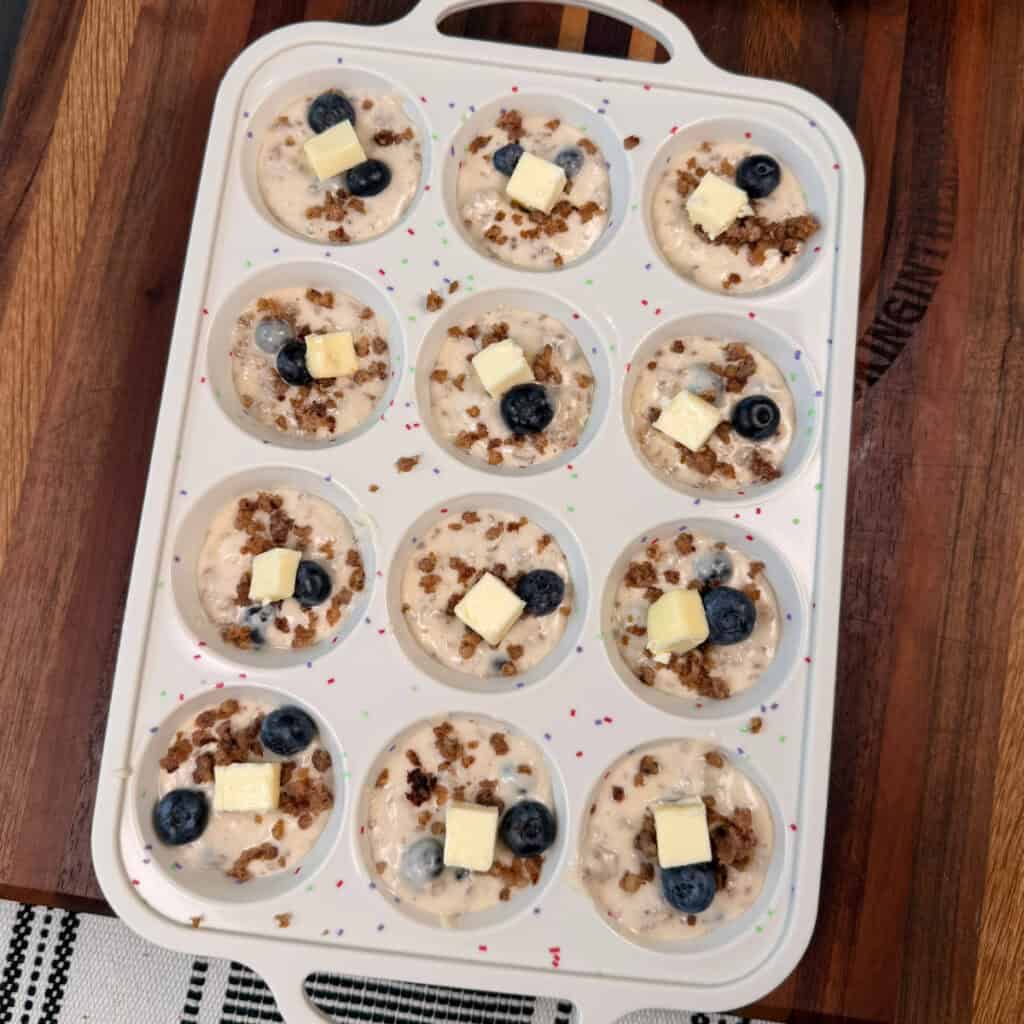 Muffin pan with uncooked pancake mix, sausage, blueberries, and topped with butter