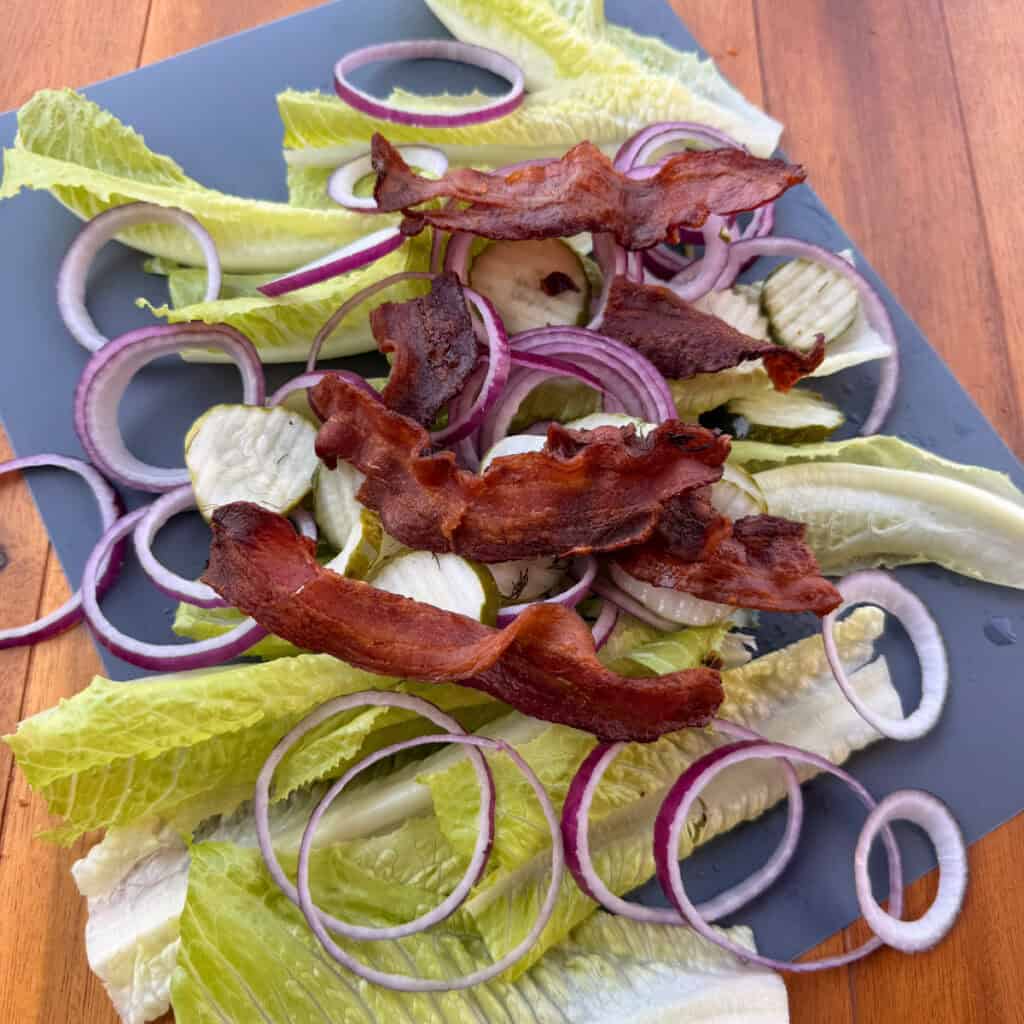 Salad, red onions. pickles, and bacon on a cutting board