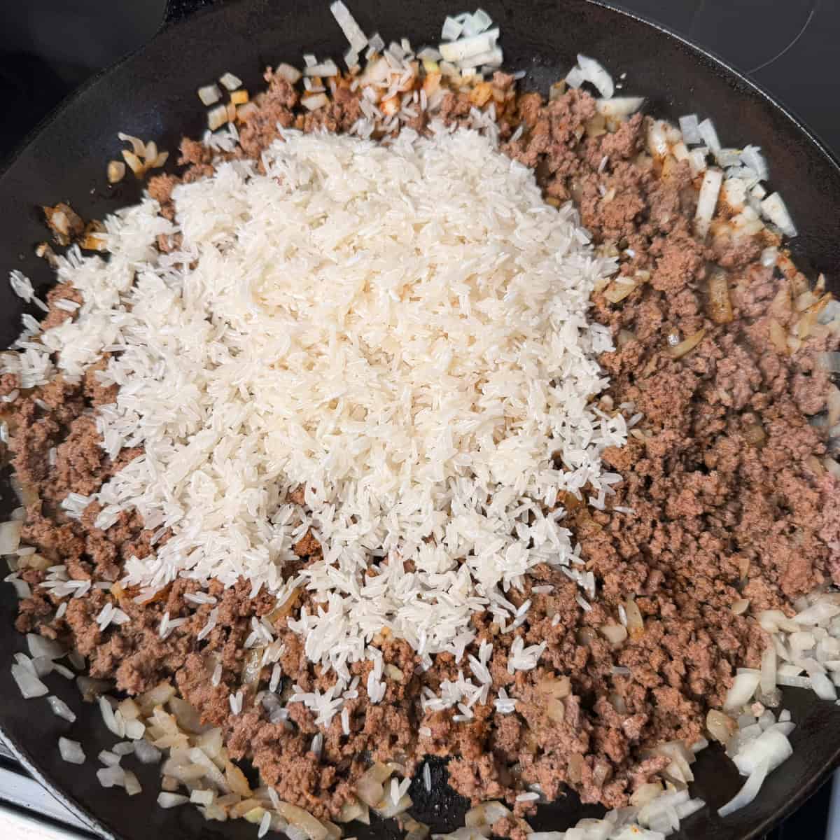Cooked ground beef and onions with rinsed Jasmine rice in cast iron skillet