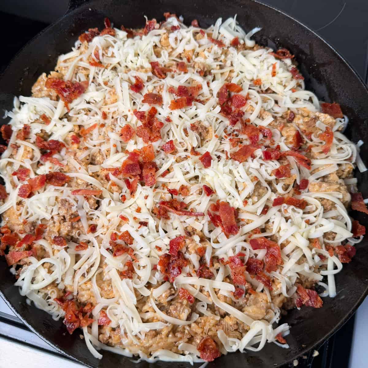 Cooked beef and rice topped with shredded mozzarella cheese and bacon in cast iron skillet